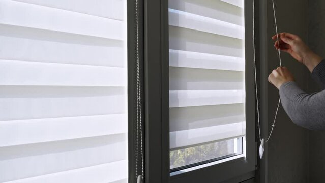 a woman opens the fabric roller blinds with day-night effect on window.