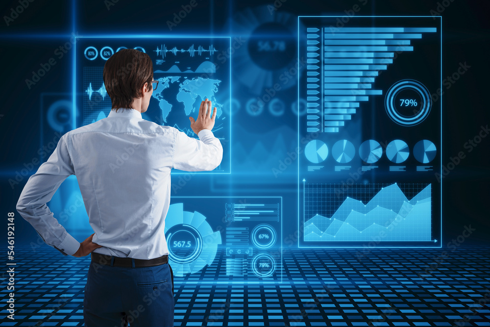 Poster Back view of attractive young man using abstract glowing blue business charts and graphs hologram on dark background. Innovation, future and fintech concept.