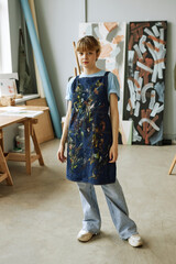 Young pretty woman in casualwear and apron standing against her workplace and paintings in studio of arts and looking at camera