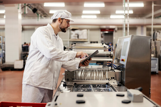 A food factory supervisor assesses the quality of the meat while typing on a tablet and looking at the packing machine.