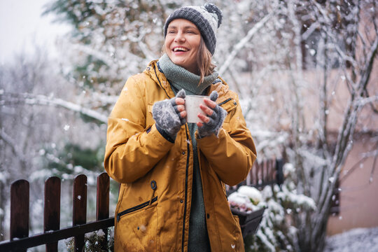 Young happy adult caucasian woman smiling in hat and yellow jacket enjoying tea in a mug with snowfall on the terrace