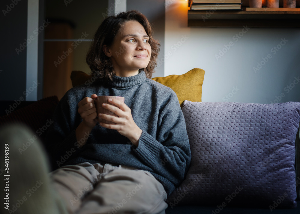 Wall mural young happy relaxed woman in warm sweater holding mug of coffee while sitting on sofa at home