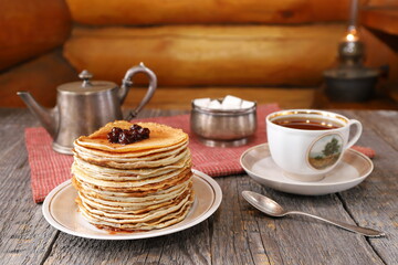 On an old wooden table against a log wall is a stack of pancakes on a ceramic plate next to vintage...