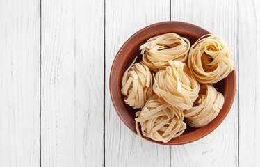 Uncooked tagliatelle pasta in ceramic bowl on white wooden background with copy space