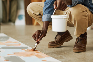 Low section of African American male artist painting artwork on canvas while squatting on rhe floor and bending over canvas in studio