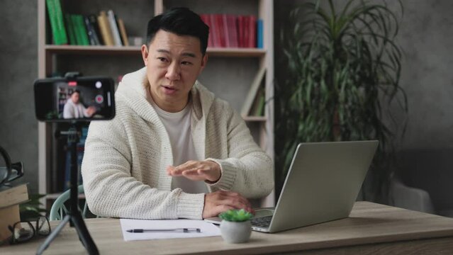 Pleasant young asian man working on laptop while recording video on his smart phone on tripod. Handsome male freelancer working remotely while staying at home.