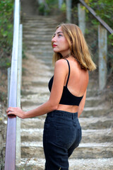 Beautiful tanned girl in top and jeans posing while standing and looking away Stairs background. Hot summer, girl beauty sexy lady, casual wear. Walking up the Stairs in the park.