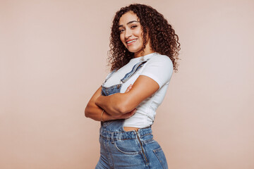 Studio portrait of cute happy smiling woman with curly hair, dressed with denim overalls and white t-shirt, with crossed arms, empty space, isolated over beige color background.