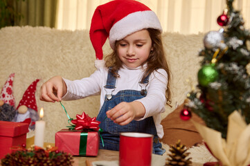 Beautiful little child girl in Santa's red hat, unpacking a cute gift box with surprise at Christmas. Winter holidays. Merry Christmas. Happy New Year. Festive mood at cozy home interior. December 25