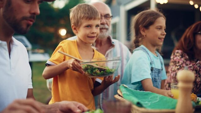 Young Boy Picking Up a Bowl of a Tasty Vegetarian Salad with Leaves and Cherry Tomatoes. Son Putting Vegan Food on a Plate to His Father. Garden Dinner with Family.