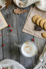 On the wooden table there is a mug of hot coffee with foam, cookies, a book, a branch of mountain ash and a warm scarf. The concept of warmth, comfort