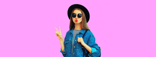 Portrait of stylish young woman blowing her red lips sending air kiss wearing black round hat, jean jacket on pink background, blank copy space for advertising text