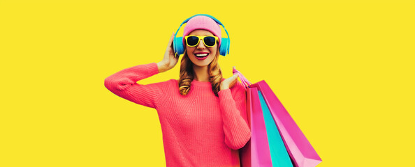 Portret van een stijlvolle, gelukkig lachende jonge vrouw die geniet van het luisteren naar muziek in een koptelefoon met kleurrijke boodschappentassen die een roze gebreide trui dragen, een muts op een gele achtergrond