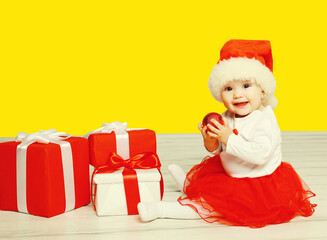 Portrait of happy smiling little child in christmas santa red hat playing with gift boxes on yellow background