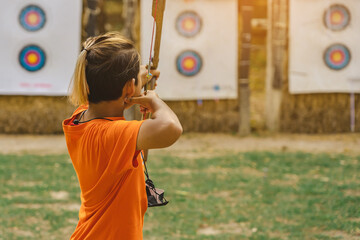 Back view of Asian boy wear face mask aims archery bow and arrow to colorful target in shooting range during training and competition. Exercise and concentration with outdoor archery. Archery for teen