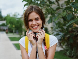 Fototapeta premium Young woman looks into the camera happiness travels on vacation with a backpack and headphones listening to music with a smile walking outside