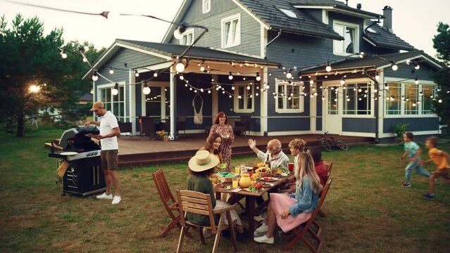 Parents, Children and Friends Gathered at a Barbecue Dinner Table Outside a Beautiful Home. Old and Young People Clapping Hands, Eating and Drinking Gorgeous Food. Garden Party in a Backyard.