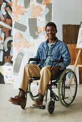 Young confident artist or craftsman in wheelchair sitting in front of camera in studio with collection of abstract paintings