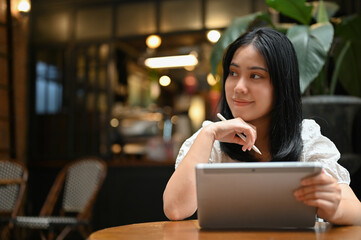 Attractive asian woman hand on chin, thinking something while using her tablet at the coffee shop.