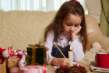 View through a blurred Christmas candle to a little child girl concentrated on writing a letter to Santa, sitting on a comfortable sofa in a cozy home interior, expecting the upcoming winter holidays