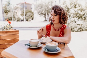 Charming woman in a restaurant, cafe on the street. She sits at the table and eats a cake with a fork. Dressed in a red sundress with white polka dots.