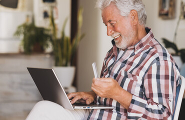 Elderly bearded man browsing by laptop enjoying black friday shopping online. Joyful and smiling senior male at home being in great mood using credit card for e-commerce purchases