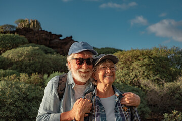 Cheerful senior caucasian couple hugging in mountain trekking enjoying nature and freedom. Smiling old retirees in hat and casual clothes among green bushes and blue sky