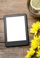 Modern e-book reader near chrysanthemums and glass of lemon water on wooden table, flat lay