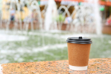 Cardboard cup with plastic lid on stone parapet near fountain outdoors, space for text. Coffee to go