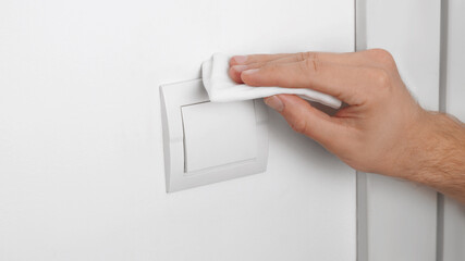 Man cleaning switch with disinfecting wipe on white wall indoors, closeup. Protective measures