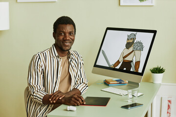 Young successful African American designer of graphic art sitting by workplace with computer monitor and picture of warrior on screen