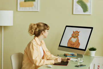 Young blond female designer or graphic artist with tablet and stylus drawing fluffy ginger cat while sitting in front of computer screen - obrazy, fototapety, plakaty