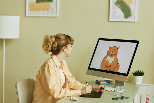 Young blond female designer or graphic artist with tablet and stylus drawing fluffy ginger cat while sitting in front of computer screen