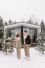 A happy couple in love in warm clothes have fun hugging and walking near a decorated country house in a snowy forest in winter outdoors. Selective focus