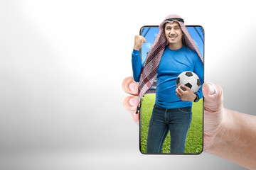 Asian man with keffiyeh standing while holding the ball with an excited expression on the football stadium