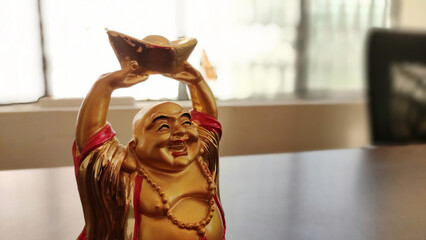 A golden Laughing Buddha statue holding a gold ingot with his hands upright. The statue is smiling broadly and wearing a red robe with a beaded necklace.