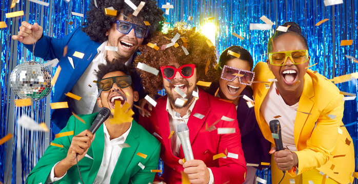 Happy Diverse Friends Having Fun At Karaoke Night. Joyful Excited People In Green, Blue, Yellow And Red Suits, Crazy Hair Style Wigs And Vintage Sunglasses Singing In Microphones At Karaoke Party