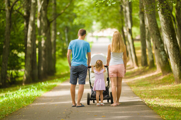 Young adult parents and little daughter pushing baby stroller and walking at tree alley of park in...