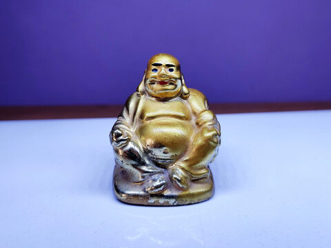A golden Laughing Buddha figurine sits on a white surface with a purple backdrop, showcasing its joyful pose, smiling face, and plump figure, symbolizing happiness, luck, and prosperity.