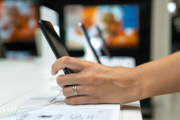 Fototapeta premium Close-up view shows the hand of a female customer choosing a black smartphone in a mobile phone store. Difficult decision, different choice