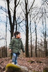 Kid standing on a rock in the woods is observing the nature and the natural environment. Child exploring the forest is staring at the tall, bare trees he's surrounded by feeling pensive and reflective