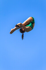 Diving Girl Athlete Unrecognizable Somersault High Action Off High Platform Against Blue Sky Into Swimming Pool.