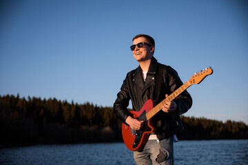 Naklejka premium a guy with an electric guitar smiling plays against the background of nature, a musician guitarist in sunglasses and a leather jacket