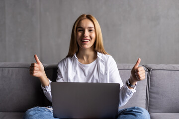 Young woman typing on her laptop on sofa and showing thumbs up