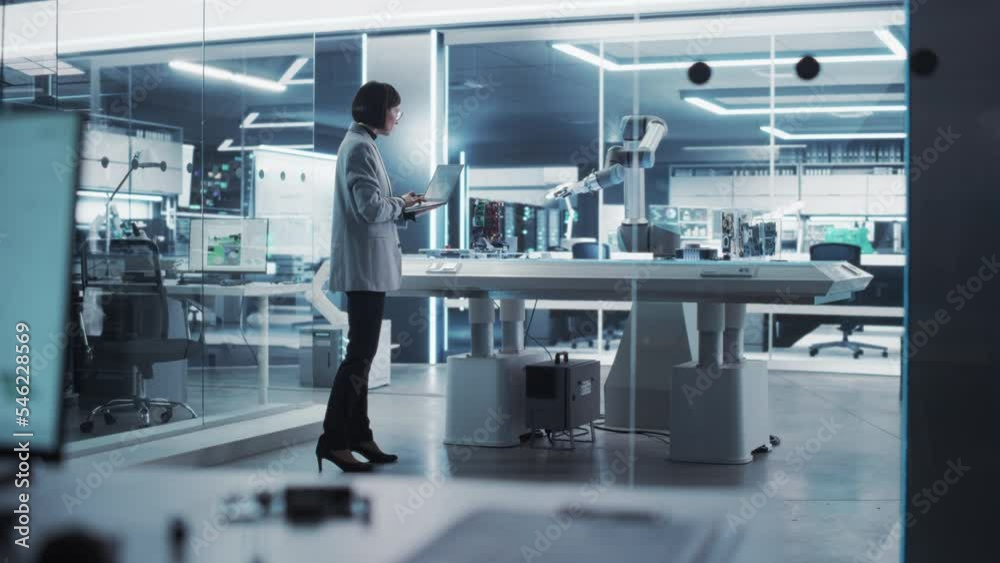 Poster Industrial Robotics Engineer Comes to a Table With Robotic Arm in a High Tech Factory. Female Scientist Uses Laptop Computer to Manipulate and Program the Robot to Pick Up and Move a Microchip.