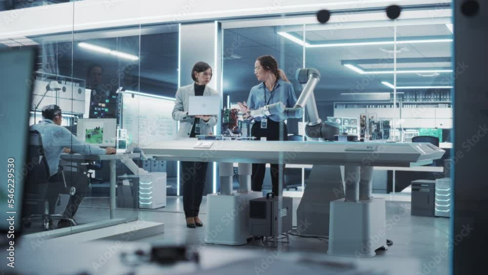 Poster Professional Multicultural Team of Two Female Engineers Using Industrial Programmable Robotic Arm in a Factory Development Workshop. Robot Hand Machine Picking Up and Holding a Microchip.