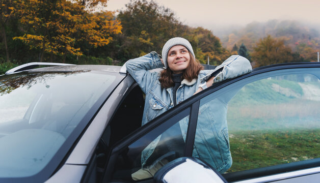 Young cheerful woman driver in a hat and jacket traveling standing next to the car in the autumn forest