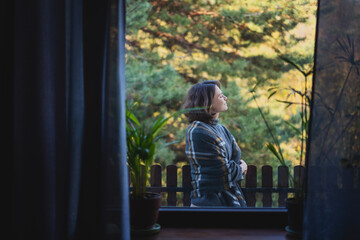 Cheerful pretty young woman 30 years old wrapped in a plaid standing on the terrace of a country house, view from the room through the window