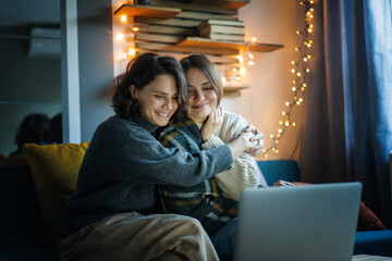 Cheerful cute lesbian couple two young women hugging in warm sweaters sitting at home on the sofa in front of a laptop