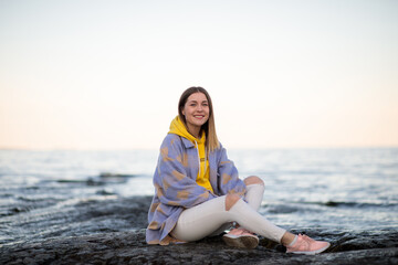 Fototapeta na wymiar a girl sits with a smile on a rocky surface in a gray coat and a yellow hoodie and white torn jeans against the backdrop of nature